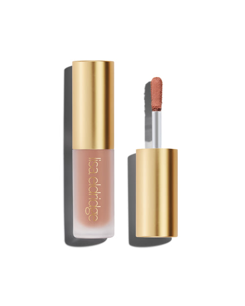 A closed bottle of Lisa Eldridge Liquid Silk Liquid Eyeshadow in peachy beige  shade Irene, with a gold cap containing an applicator wand with product on it, against a white background.