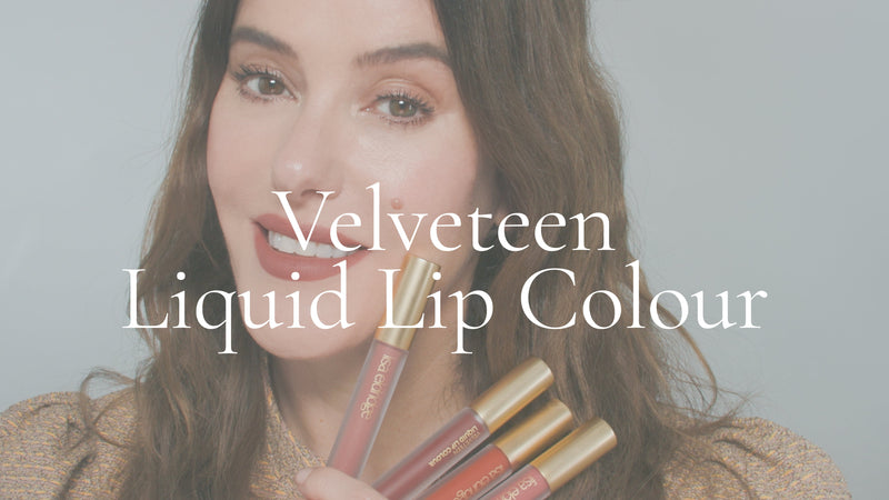 Poster image with white overlay for a video. Lisa Eldridge is in front of a grey background, holding a four Velveteen Liquid Lip Colour tubes. On top of the image is the text, “Velveteen Liquid Lip Colour”.