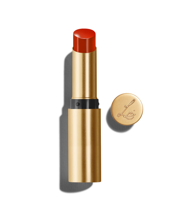 Baume Embrace in Red Curve, a bright red lip colour, with a gold-colored casing, next to the Lisa Eldridge logo-embossed lid